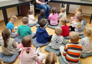 Grupa dzieci siedzi na poduszkach i słucha książki czytanej przez panią bibliotekarkę