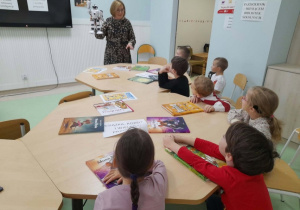 Dzieci siedzą przy stoliku, słuchają pani bibliotekarki, które pokazuje im robota.