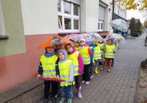 Grupa dzieci spaceruje podczas padającego deszczu. Dzieci są ubrane w odpowiedni strój i mają parasolki.