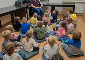 Grupa dzieci w bibliotece siedzi na poduszkach na podłodze i słucha wypowiedzi pani bibliotekarki