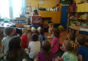Dzieci siedzą na dywanie nauczycielka pokazuje otrzymaną nagrodę – gra „Alfabet”