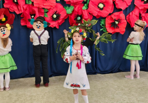 Grupa dzieci w przebraniach podczas recytacji wiersza, w tle dekoracja - duże, czerwone maki.