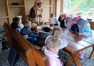 Grupa dzieci siedzi przy stole i słuchaja pani, która opowiada o ptakach.