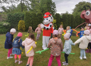 Dzieci trzymają kolorowe balony, otaczają dużą, chodzącą maskotkę psa - strażaka.