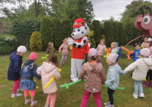Dzieci trzymają kolorowe balony, otaczają dużą, chodzącą maskotkę psa - strażaka.