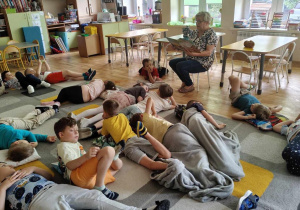 Pani nauczycielka czyta dzieciom książkę, dzieci odpoczywają na dywanie.