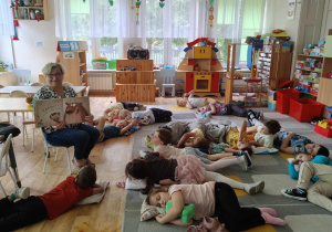 Pani nauczycielka czyta dzieciom książkę, dzieci odpoczywają na dywanie.