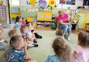 Pani nauczycielka czyta dzieciom książkę, dzieci siedzą na dywanie.