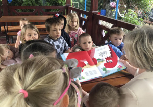 Pani nauczycielka czyta dzieciom książkę, wszyscy zgromadzeni są w ogrodowej altanie.