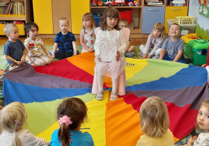 Dzieci siedzą wokół chusty animacyjnej, dziewczynka jubilatka siedzi w środku kręgu na krzesełku.