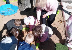 Dzieci w piaskownicy wraz z panią pedagog szukają muszli.