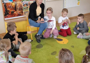 Dzieci siedzą na dywanie i słuchają czytanej książki.