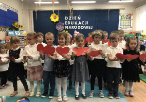 Grupa dzieci (w eleganckich, galowych strojach) podczas występu z okazji Dnia Nauczyciela, dzieci trzymają czerwone serduszka w dłoniach.