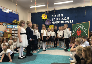 Grupa dzieci (w eleganckich, galowych strojach) podczas prezentacji skeczu z okazji Dnia Nauczyciela.