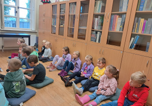 Grupa dzieci siedzi na poduszkach na podłodze i słucha wypowiedzi pani bibliotekarki
