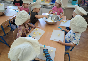 Grupa dzieci przy stolikach na deseczkach do krojenia, kroi owoce na sałatkę owocową i przekłada do dużej miski stojącej na środku stolika.