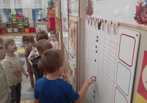 Grupka dzieci stoi przy tablicy z magnezem w ręku i odszukuje swój znaczek.