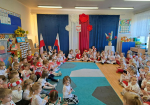 Dzieci (w biało-czerwonych strojach) zebrane w patriotycznie udekorowanej sali przedszkolnej.