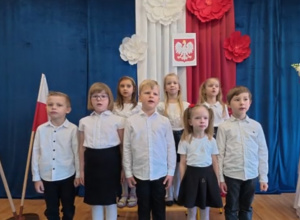 Grupa dzieci w galowych strojach na biało-czerwonym tle.