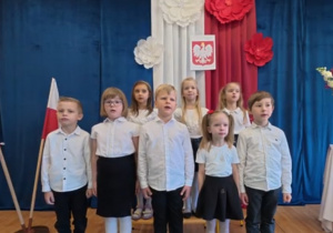 Grupa dzieci w galowych strojach na biało-czerwonym tle.