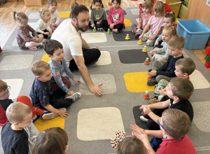 Dzieci siedzą na dywanie w kole i grają na dzwonkach w rytm muzyki.