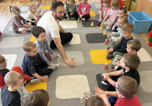 Dzieci siedzą na dywanie w kole i grają na dzwonkach w rytm muzyki.