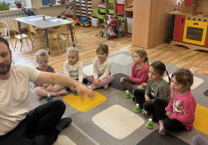 Dzieci siedzą na dywanie w kole i grają na dzwonkach w rytm muzyki.