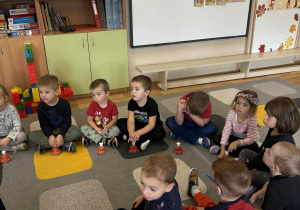 Dzieci siedzą na dywanie w kole i grają na dzwonkach w rytm muzyki.