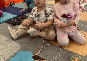 Dzieci siedzą na dywanie przed tekturowymi sylwetkami słońca i chmurek, do których dołączone są sznurki.