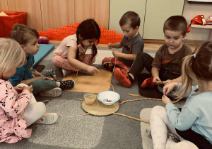 Dzieci siedzą na dywanie przed tekturowymi sylwetkami słońca i chmurek, do których dołączone są sznurki.