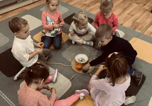 Dzieci siedzą na dywanie przed tekturowymi sylwetkami słońca i chmurek, do których dołączone są sznurki.