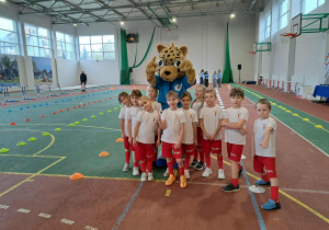 Dzieci w sportowych strojach pozują do zdjęcia z maskotką - dużym lampartem.