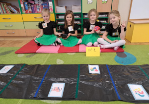 Cztery dziewczynki siedzą na materacu, jedna z nich trzyma dużą, czerwoną kostkę do gry.