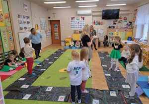 Dzieci stoją na dużej planszy do gry rozłożonej na dywanie.