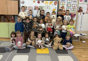 Dzieci stoją na dywanie w sali i prezentują swoje misie