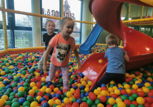 Troje dzieci bawi się w suchym basenie z kolorowymi piłeczkami.