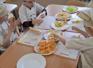 Dzieci siedzą przy stolikach i kroją owoce.