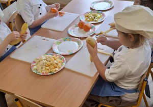 Dzieci siedzą przy stolikach i obierają owoce.