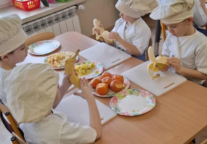 Dzieci siedzą przy stolikach i obierają owoce.