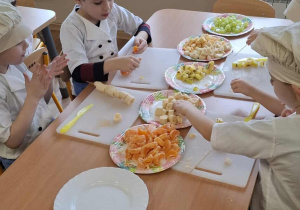 Dzieci siedzą przy stolikach i obierają owoce.