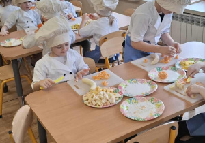 Dzieci siedzą przy stolikach i kroją owoce.