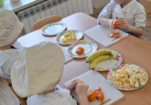 Dzieci siedzą przy stolikach i obierają owoce.