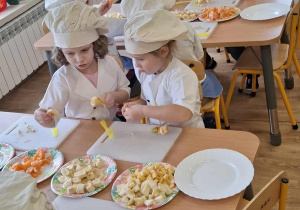 Dzieci siedzą przy stolikach i nadziewają owoce na wykałaczki.