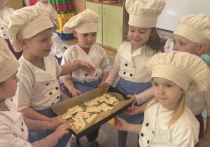 Grupka dzieci trzyma w rękach blachę ze swoimi ciasteczkami naszykowanymi do pieczenia.