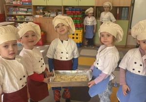 Dzieci stoją na dywanie trzymając ciastka na metalowej blasze.