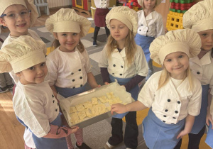 Dzieci stoją na dywanie trzymając ciastka na metalowej blasze.