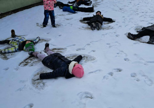 Pięcioro dzieci leży na śniegu i robi tzw. „aniołka” a troje dzieci stoi.