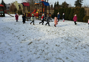Grupa dzieci biegnie „gęsiego” po śniegu wokół drewnianego zestawu do zabawy.