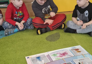 Dzieci siedzą ba dywanie, chłopiec ogląda swoją dłoń przez lupę.