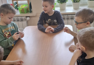 Dzieci siedzą przy stoliku, chłopiec przykładam magnes do kubeczka ze spinaczem biurowym.